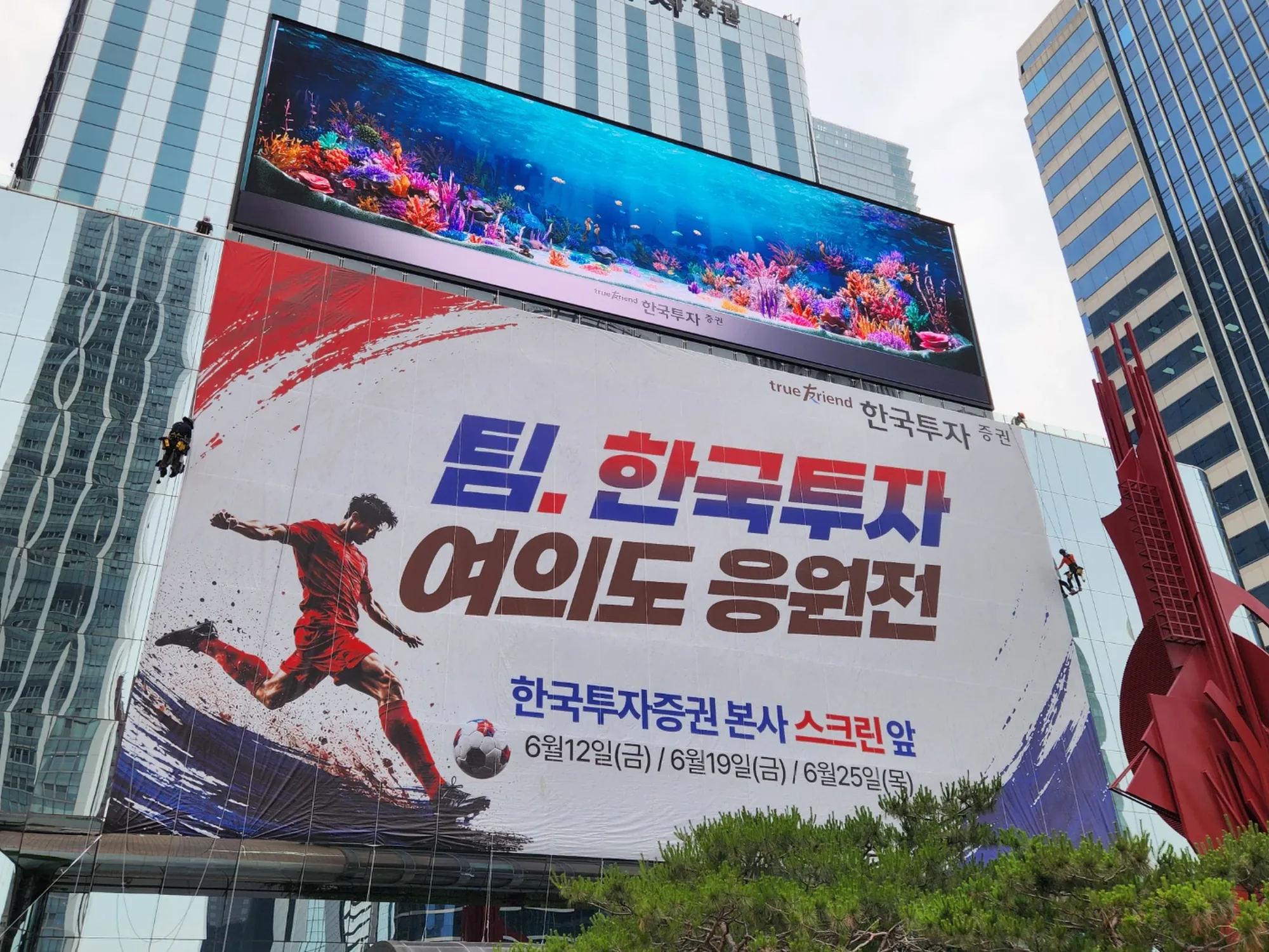 건물 외벽에 설치된 대형 응원 현수막과 중앙 붉은 조형물, 로프접근 작업자가 양옆에서 마무리 작업 중인 모습